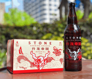 A 12oz bottle of Stone Soaring Dragon IPA next to a six pack of Stone Soaring Dragon IPA cans on a table